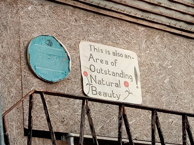 Two home-made signs, one mimicking a blue plaque, another saying "This is also an Area of Outstanding Natural Beauty"