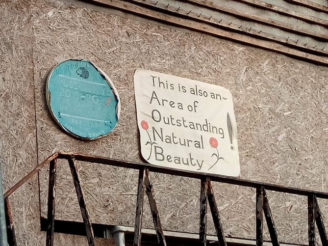 Two home-made signs, one mimicking a blue plaque, another saying "This is also an Area of Outstanding Natural Beauty"
