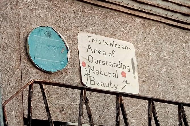 Two home-made signs, one mimicking a blue plaque, another saying "This is also an Area of Outstanding Natural Beauty"